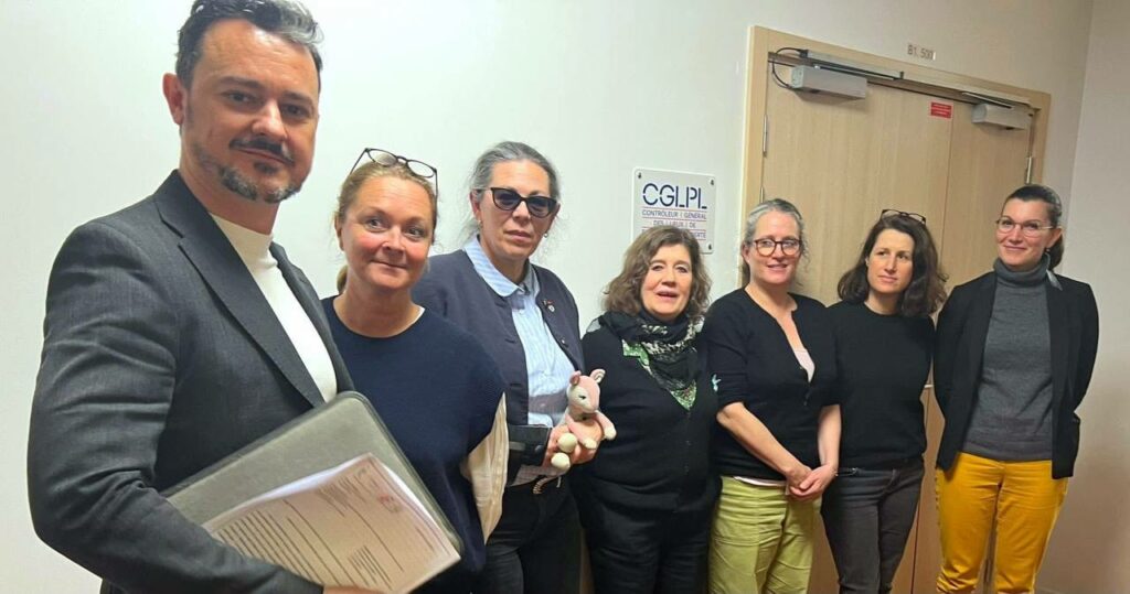 Six personnes posent dans un couloir devant une porte portant le sigle "CGLPL". L'une tient un dossier, une autre une peluche, dans un contexte de rencontre institutionnelle.