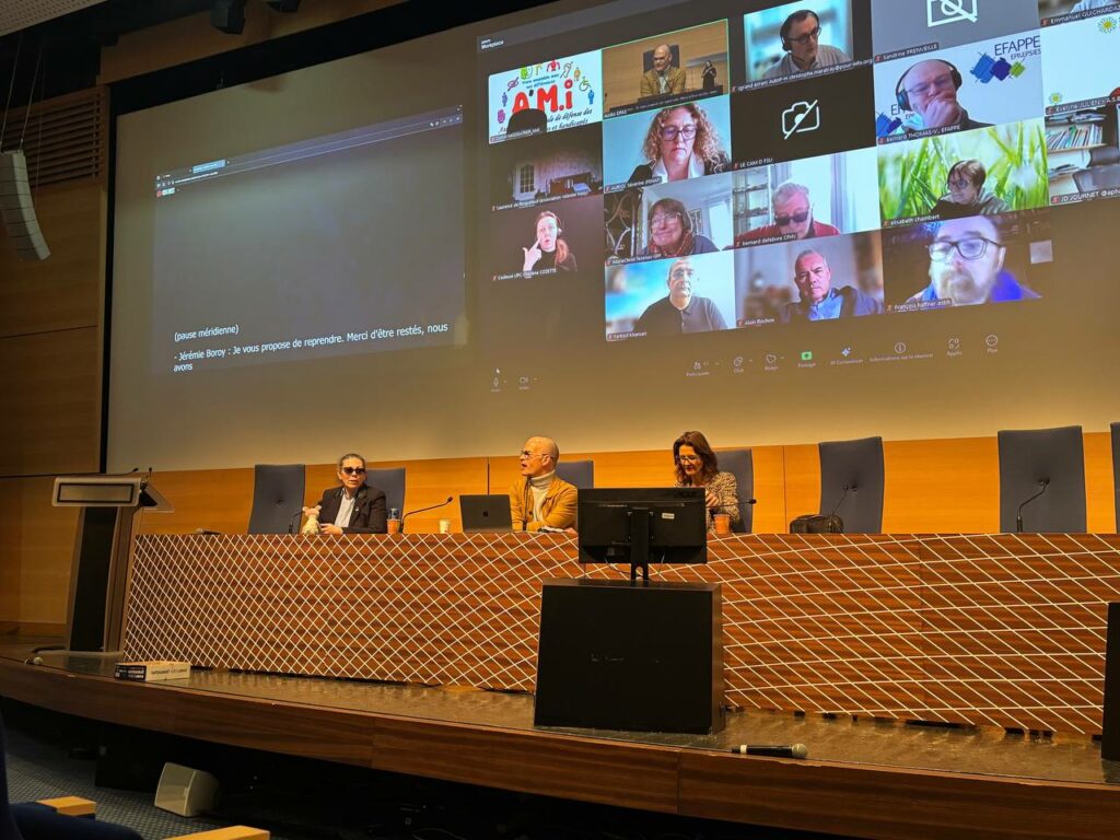 Salle de conférence avec trois intervenants assis derrière une grande table sur scène.
Un écran géant projette une visioconférence affichant plusieurs participants à distance.
Des micros et ordinateurs sont disposés devant les intervenants.
L’ambiance est institutionnelle, avec un pupitre à gauche et un éclairage sobre.
Un micro repose au sol devant l’estrade.
